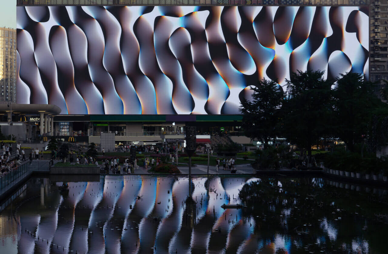 Picture of the plaza with a pond reflecting the screen showing the artwork, as people casually walk and move through the space.
