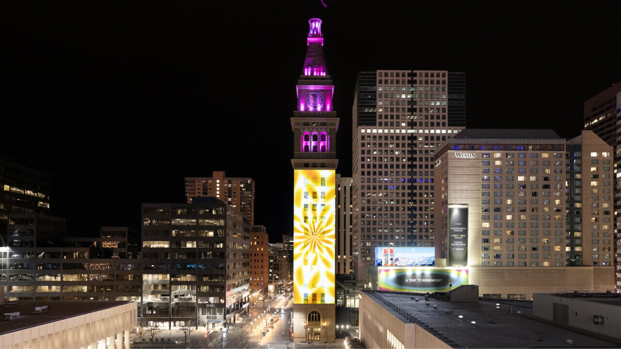 Projection of digital artwork displayed on a tall clock tower at night, blending vibrant colors and patterns with the architectural structure.