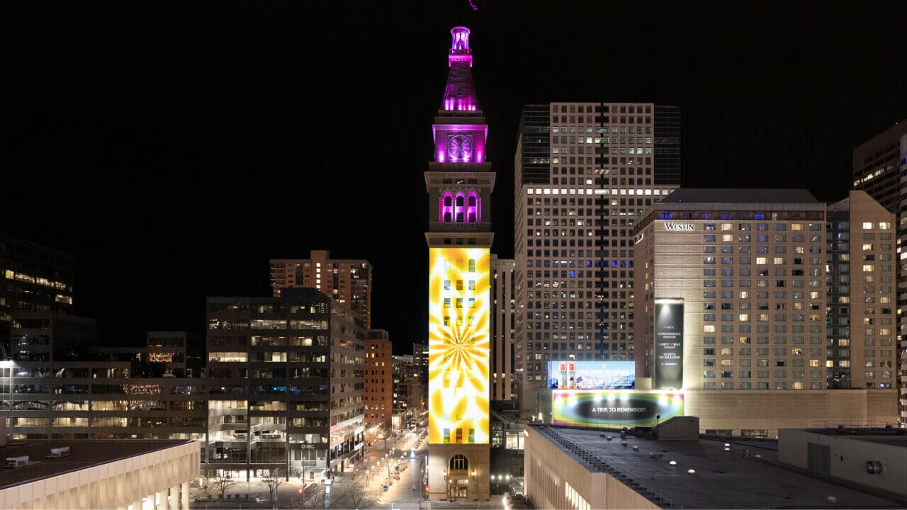 Projection of digital artwork displayed on a tall clock tower at night, blending vibrant colors and patterns with the architectural structure.