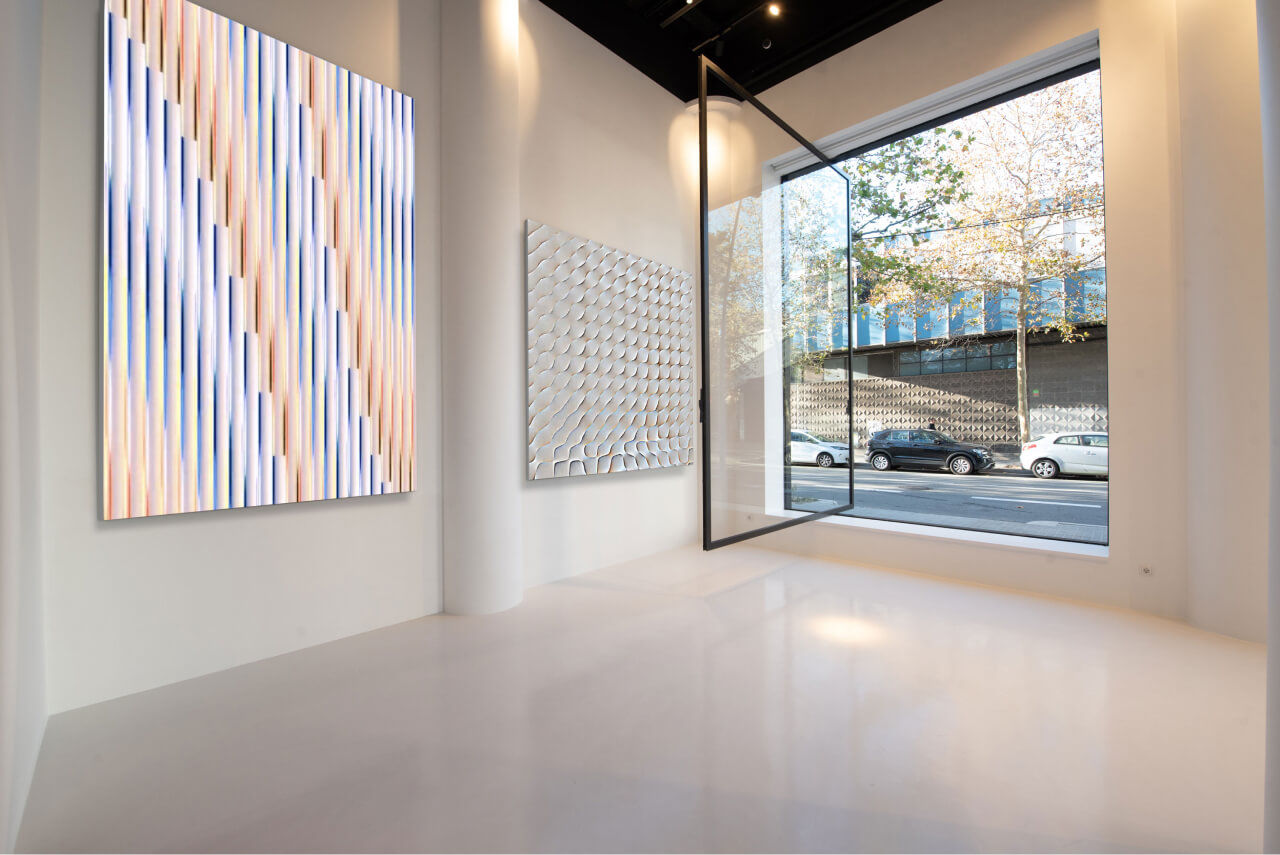 Wide view of a modern white gallery with LED screens displaying the artist's work, featuring a large window open to the street.